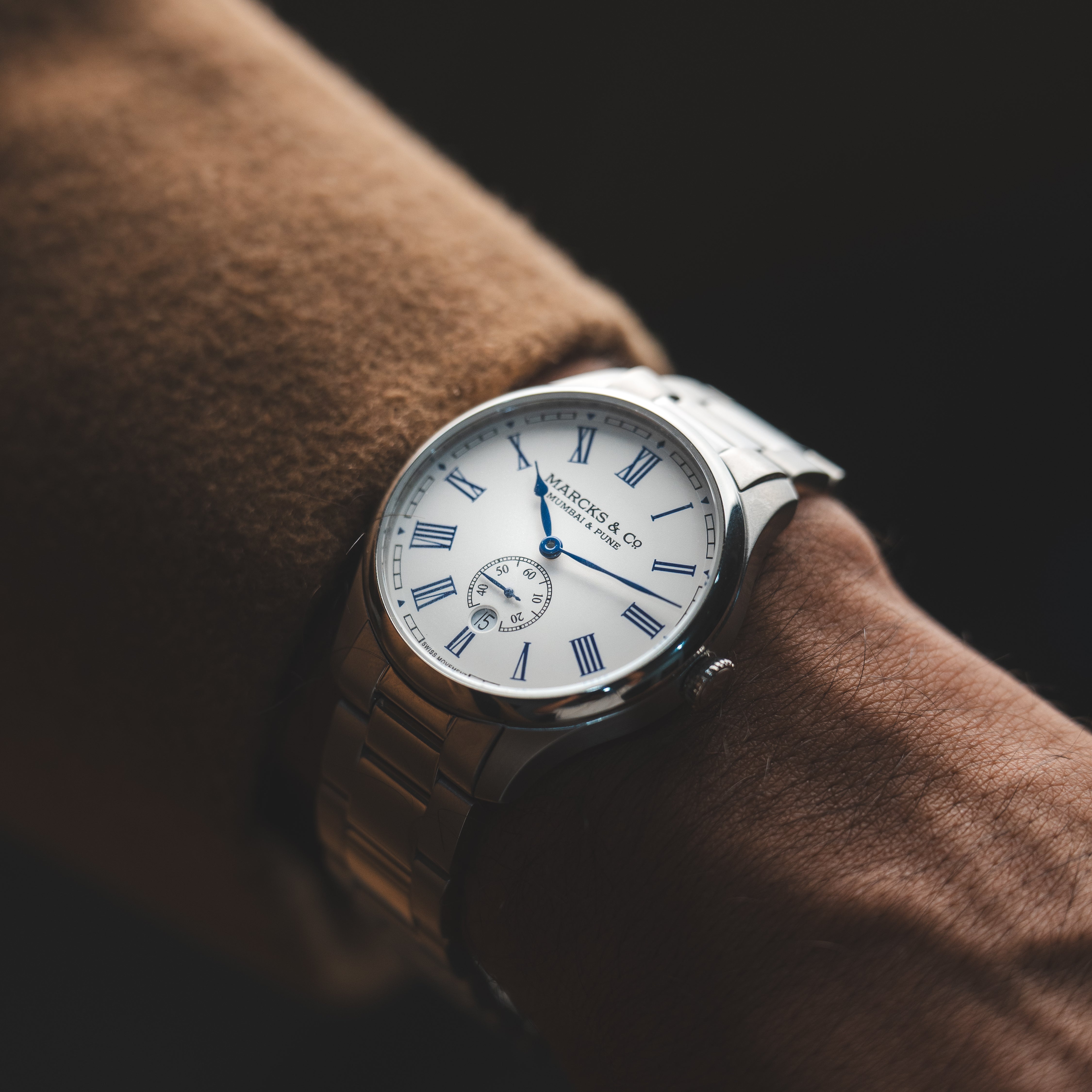 Silver watch with a white face and blue accents on a brown wrist against a dark background